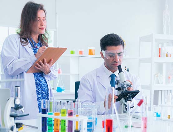Two scientists working in a lab. One is looking in a microscope and one is taking notes.