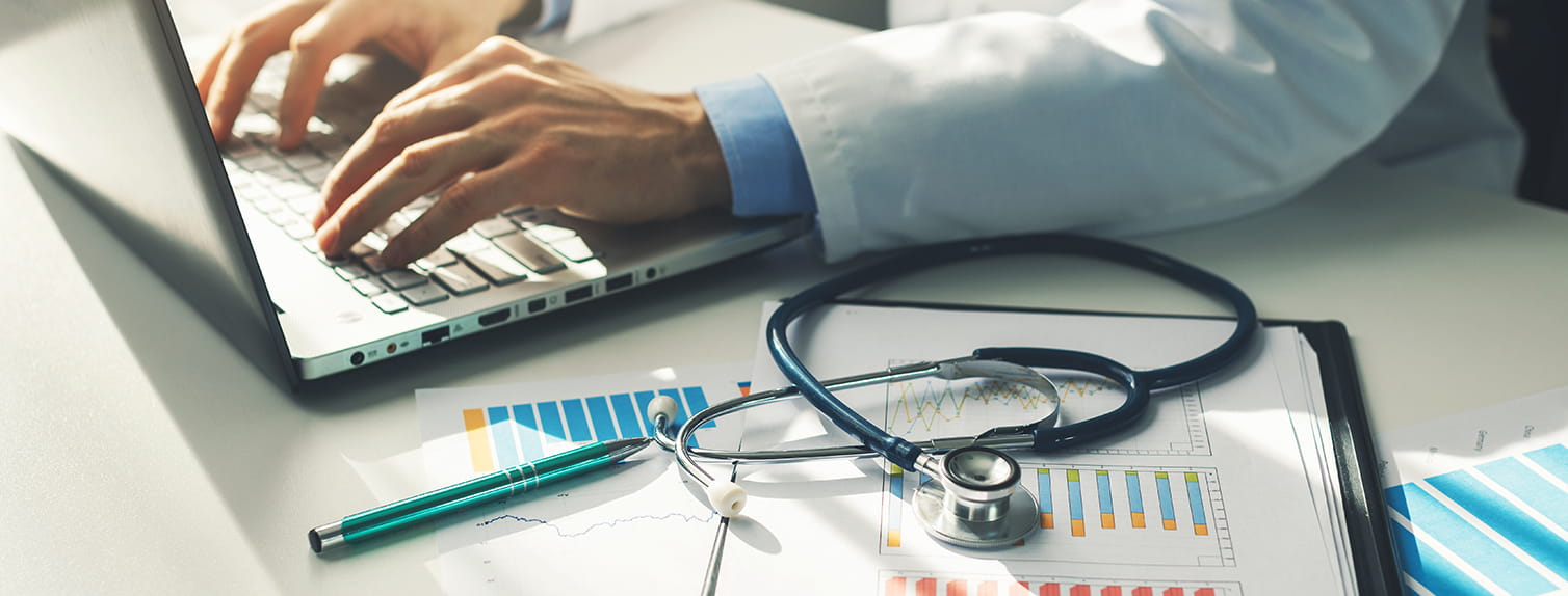 doctor sitting at desk typing on computer with charts and stethoscope 
